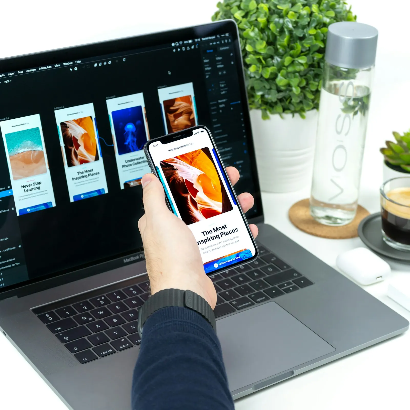 Hand holding a phone with a mobile app open, with the app's screen designs on a laptop behind